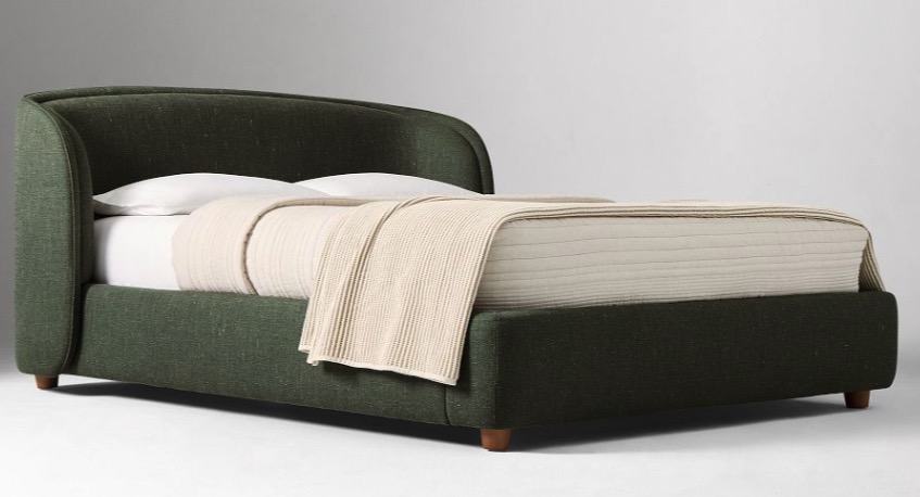 An upholstered bed in deep green tweed with a curved, winged headboard, made up with white pillows and an oatmeal throw
