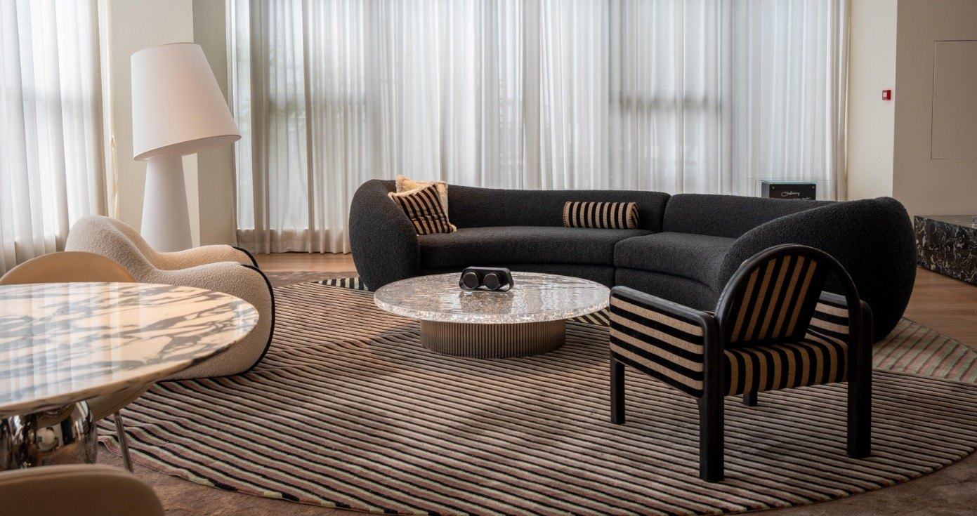 A living room with a curved charcoal bouclé sofa, a striped lounge chair, a glass coffee table and a round striped rug, by tall sheer curtains
