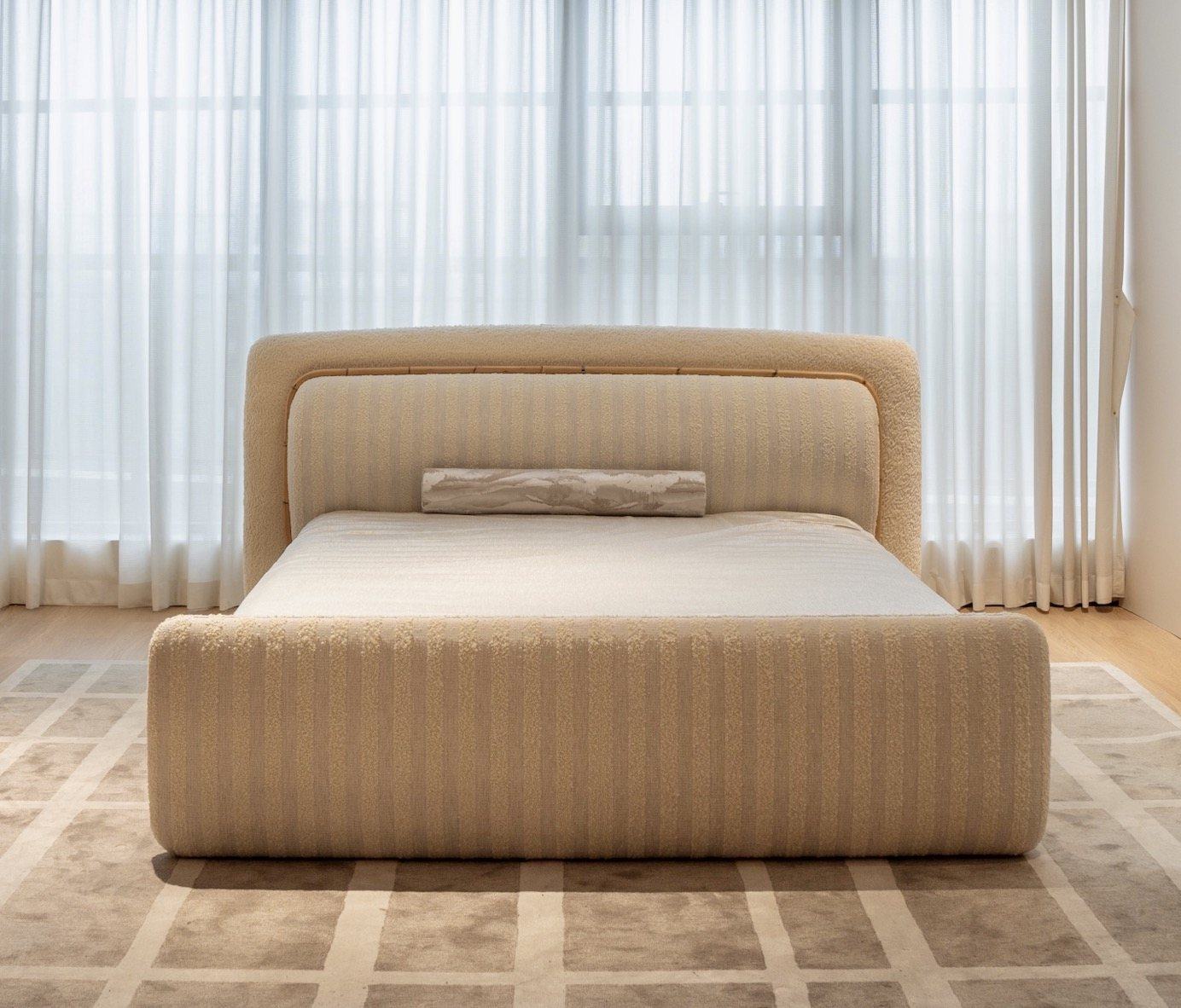 An ivory bouclé bed with a striped upholstered headboard and footboard in front of sheer curtains